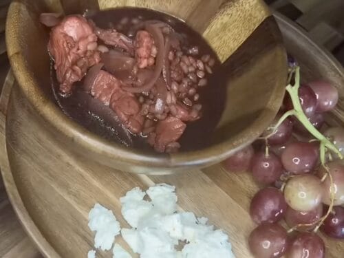 Bowl of dark reddish broth with meat chunks, beside diced white onions and fresh grapes on wooden board